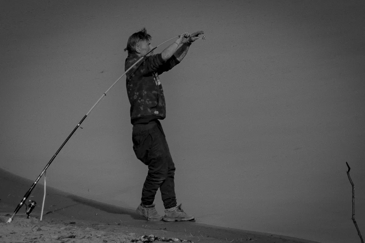 Person fishing on a sandy bank with a bent rod against an empty horizon